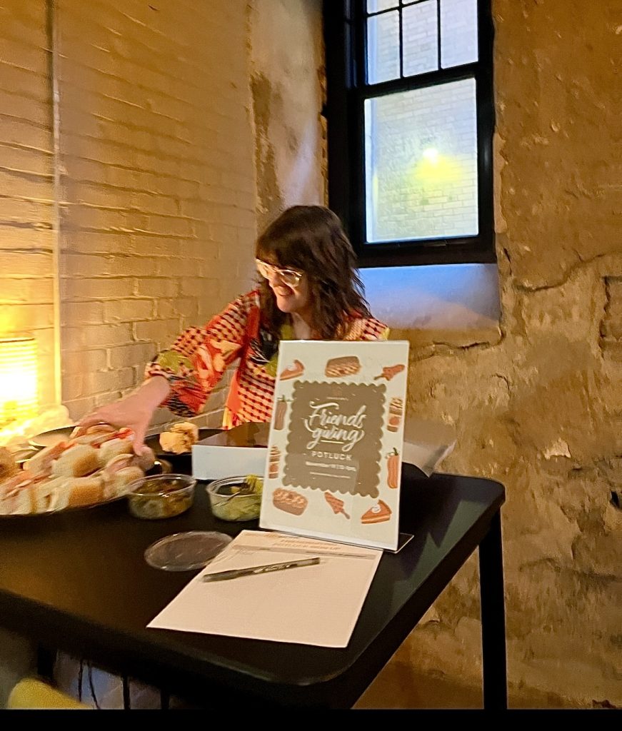 Person smiling and arranging food at a Friendsgiving potluck table inside Groundwork coworking space, with a warm brick wall, soft lighting, and a festive Friendsgiving sign in view.