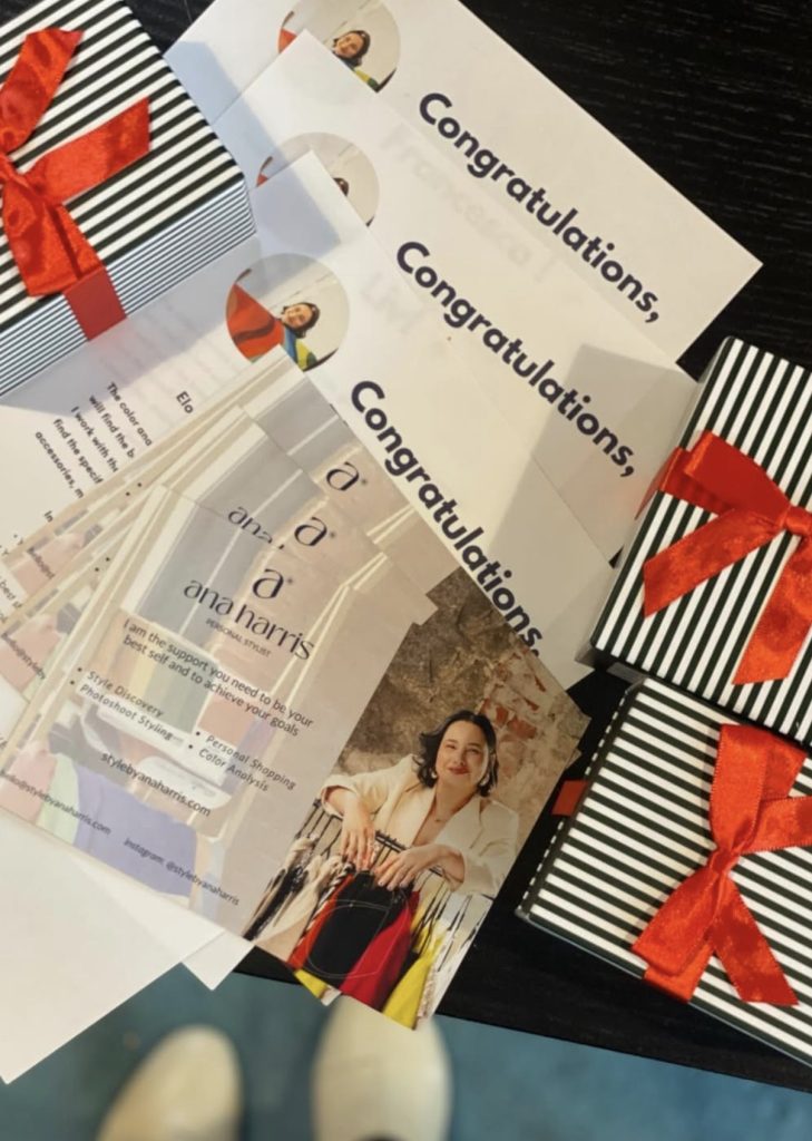 Gift certificates and business cards for Style by Ana Harris are displayed alongside black-and-white striped gift boxes tied with red ribbons. The cards feature Ana Harris smiling while holding a clothing rack, promoting personal styling, shopping support, and color analysis services. The scene conveys an elegant, celebratory presentation with the word “Congratulations” visible on several certificates.
