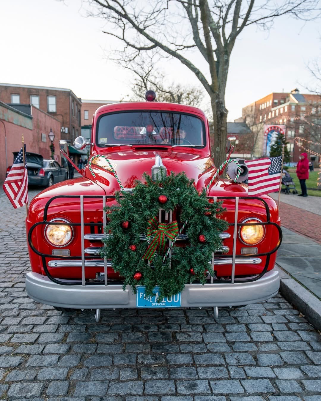 A bright red vintage fire truck decorated for the holidays sits on a cobblestone street with headlights glowing. A large Christmas wreath with red ornaments hangs on the front grille, and two American flags are mounted on either side of the hood. Buildings, trees, and a festive street scene are visible in the background.