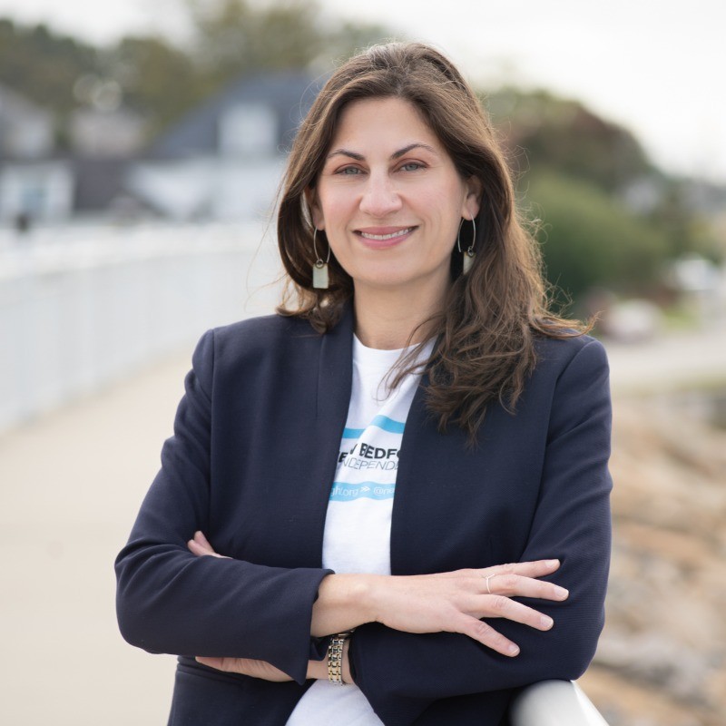 The headshot of a woman named Colleen Trahan of Whale rock consulting. She is standing outdoors wearing a blazer smiling at the camera with her arms folded.