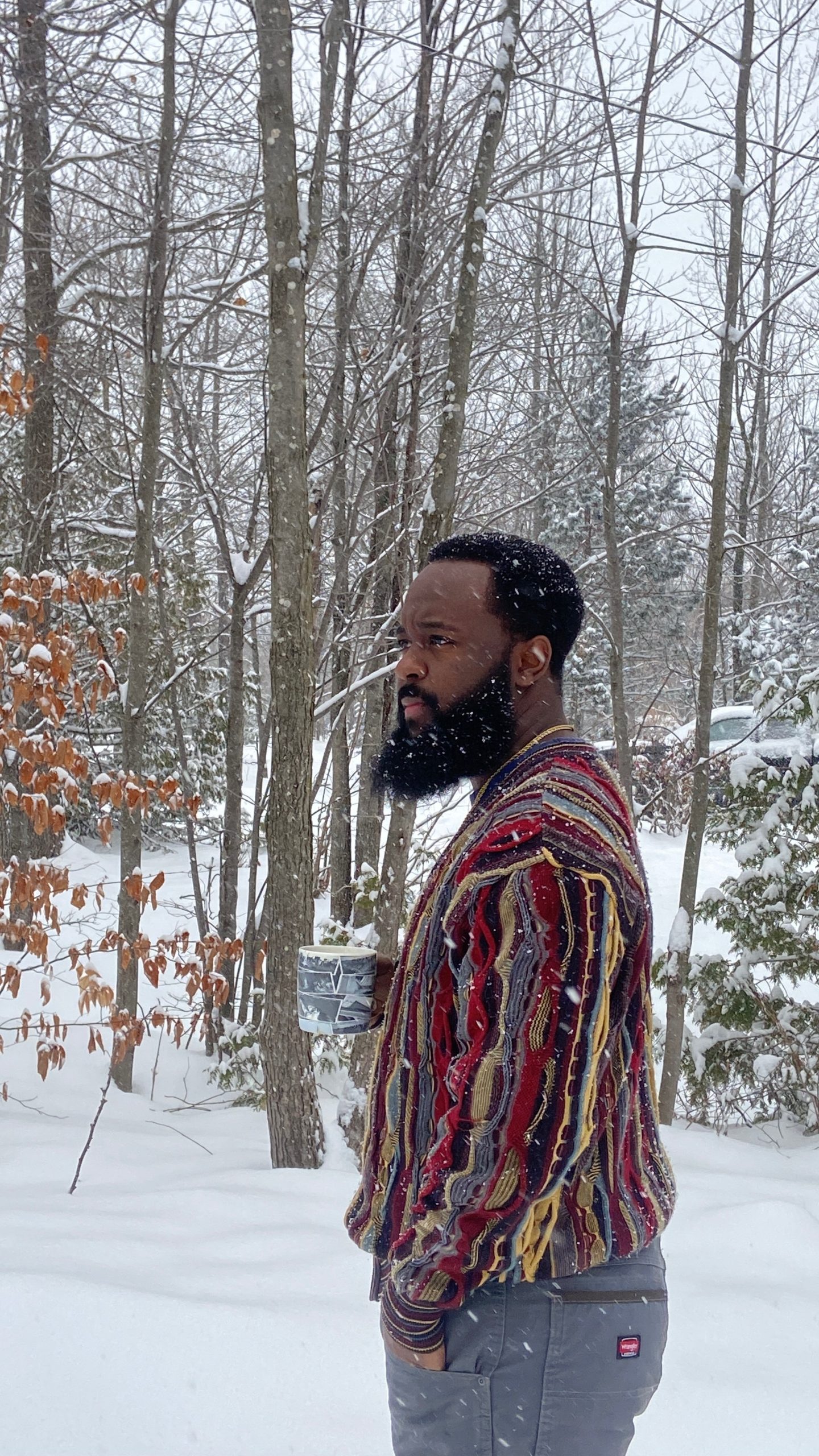 Garnelli Marcel standing outdoors in a snowy New England forest holding a mug, surrounded by trees in winter