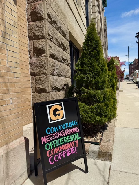 Sidewalk view outside Groundwork Fall River featuring a black sandwich board sign advertising coworking, meeting rooms, office rentals, community, and coffee, with evergreen shrubs lining the building entrance.
